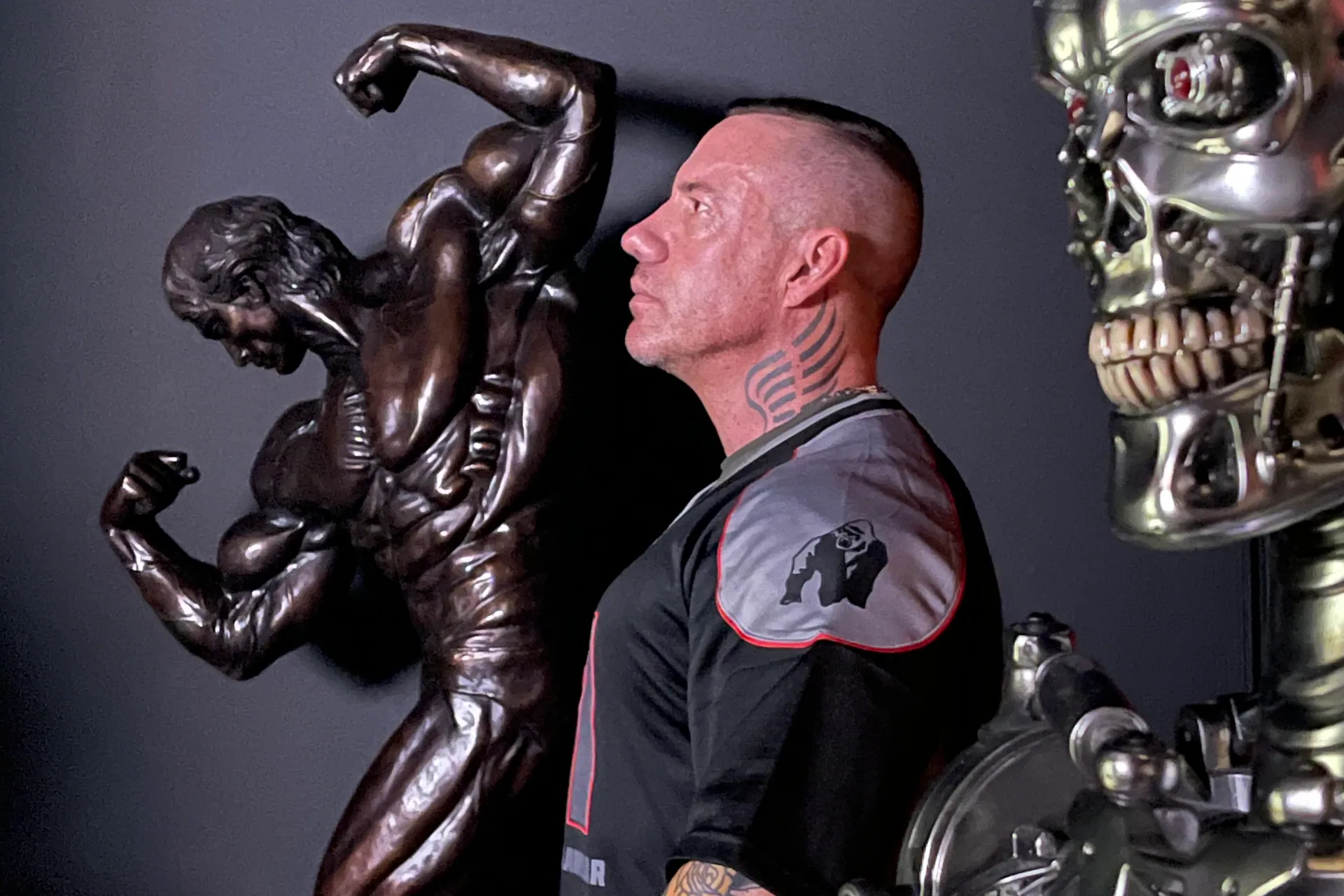 Marc Berbet in profile between a bronze classical bodybuilder statue and a chrome T-800 Terminator endoskeleton